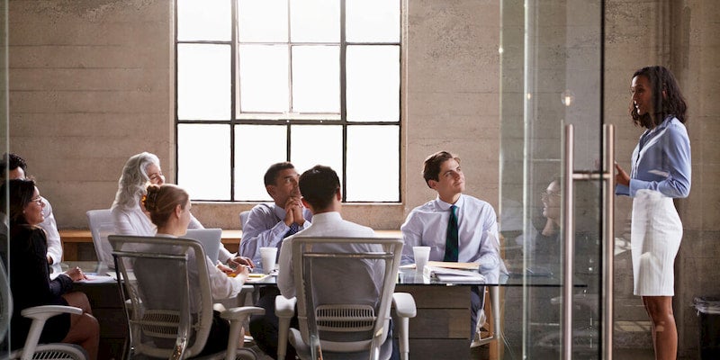 Glass doors open to a meeting room, a consultant is giving a presentation and a group of professionals are watching and smiling.