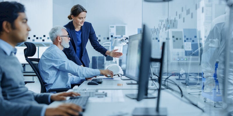 A business consultant stands next to a worker at a desk. She is explaining something on his computer. 