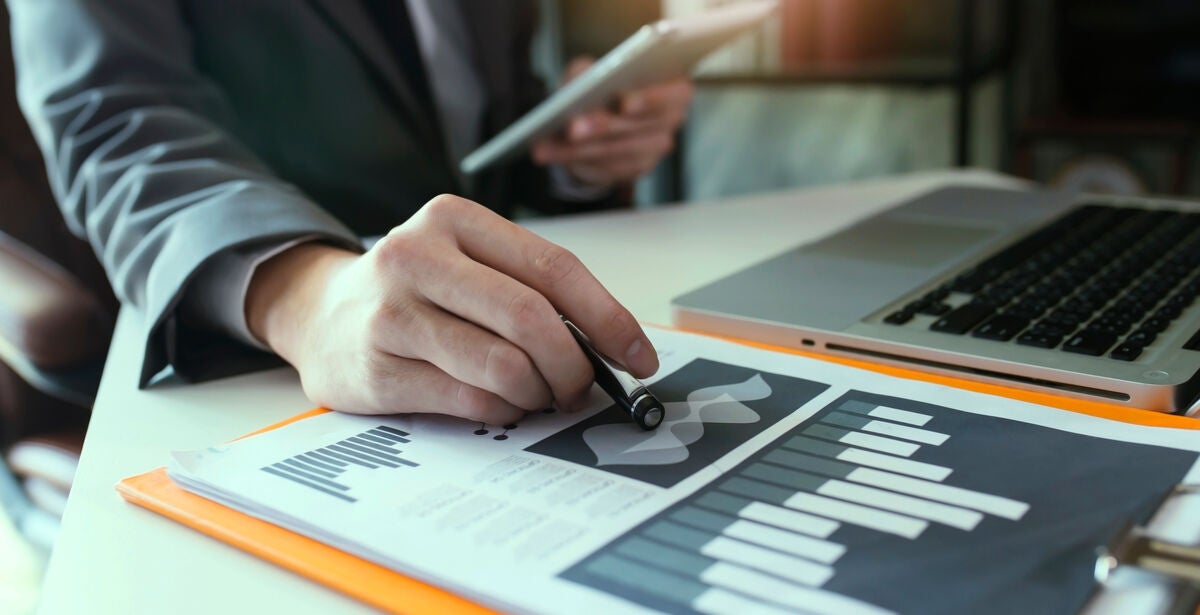 A desk with a graph and a hand holding a pen in the foreground, a laptop and person holding a clipboard in the background.