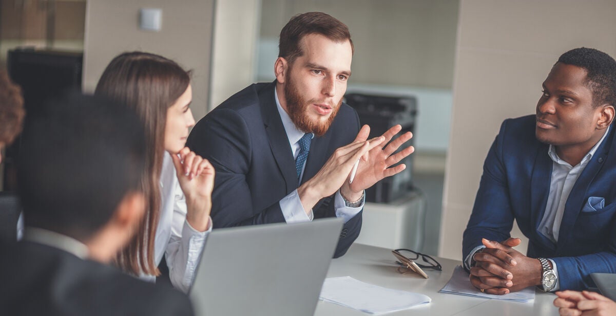 A group of professionals sit around a table, listening intently to a business consultant talking.