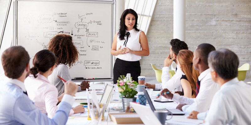 A business consultant stands in front of a whiteboard, making a presentation to a group of people.