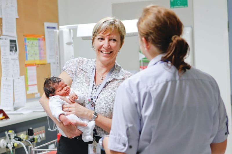 A nurse is holding a baby and smiling while talking to another nurse.