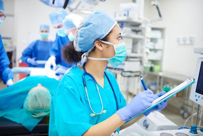 A nurse is in an operating theatre, looking at a machine and taking notes.
