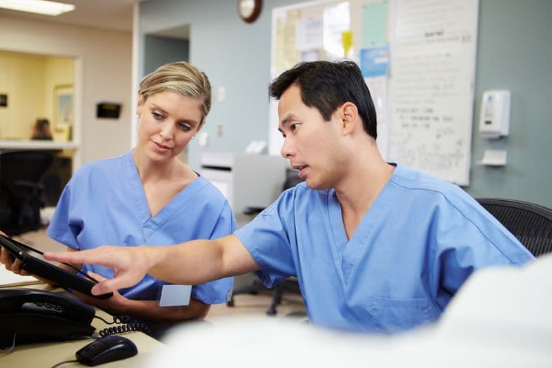 Two nurses are looking at a tablet. One is pointing at it and explaining something to the other.