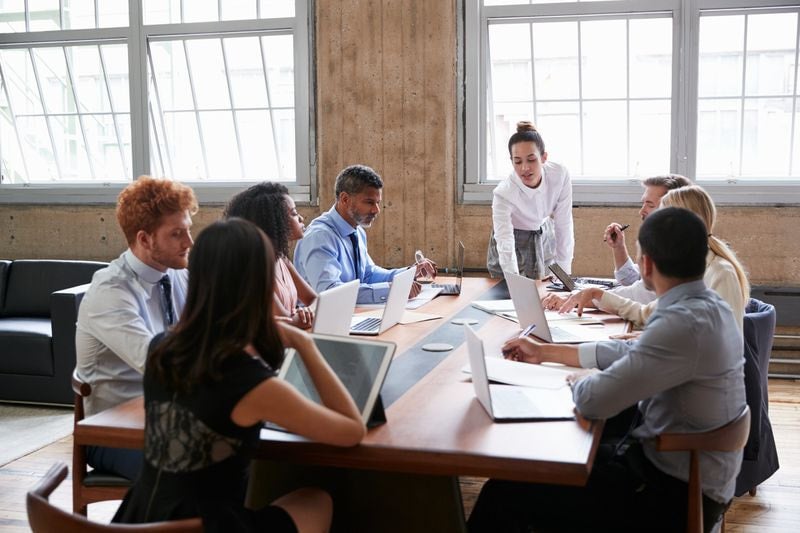 A business consultant is leading a meeting, she is talking and a group of people are listening and reading notes.