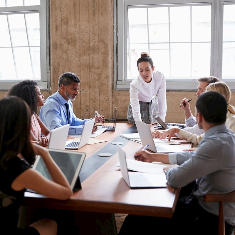 A business consultant is leading a meeting, she is talking and a group of people are listening and reading notes.