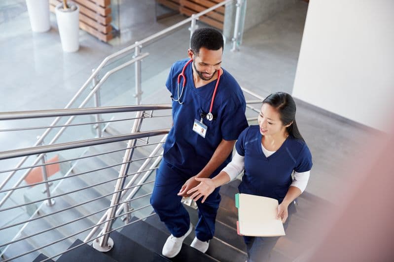Two nurses walk up a flight of stairs in a hospital, talking and smiling.