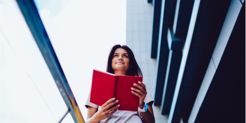 Young girl reading a book while working through university grounds