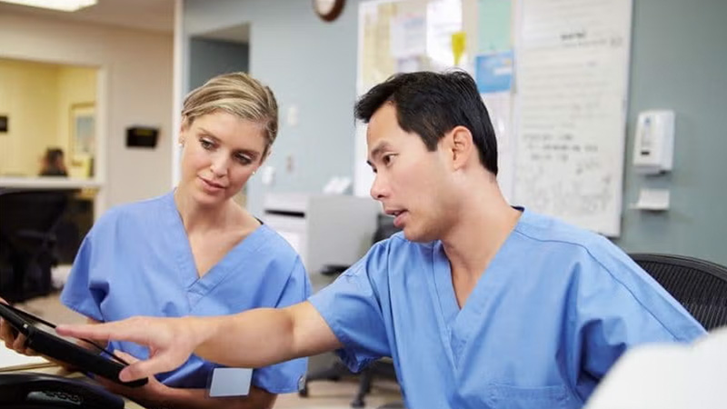 Two medical professionals in scrubs discussing on a tablet in an office setting.