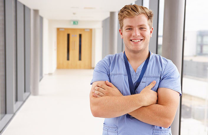 Male nurse in blue scrubs smiling with arms crossed in a hospital hallway.