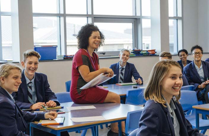 A teacher engages with smiling high school students in a classroom.