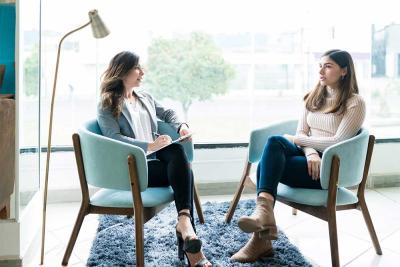 Two women engaged in conversation while seated in chairs, sharing thoughts and laughter in a relaxed setting.