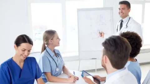 A nurse leader presents information to a group of nurses in a meeting room.